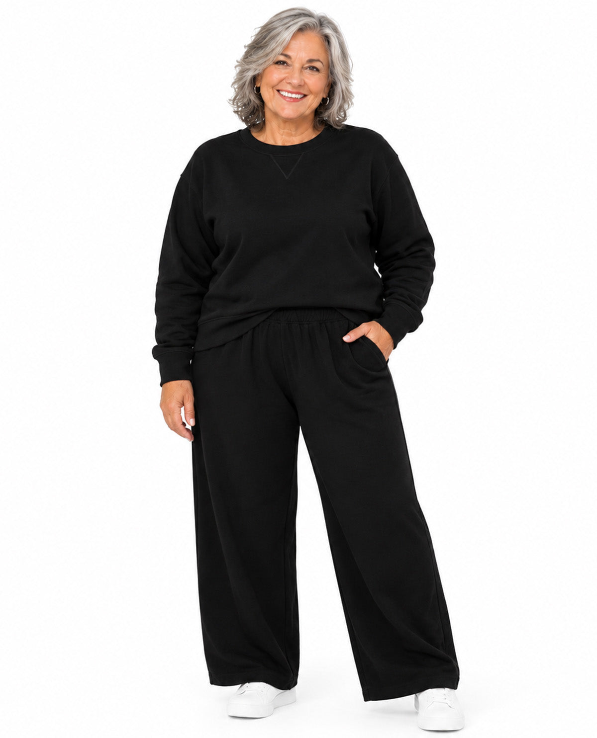 A smiling woman with gray hair stands against a plain white background wearing a black long-sleeve sweatshirt and matching black wide-leg pants, paired with white sneakers.