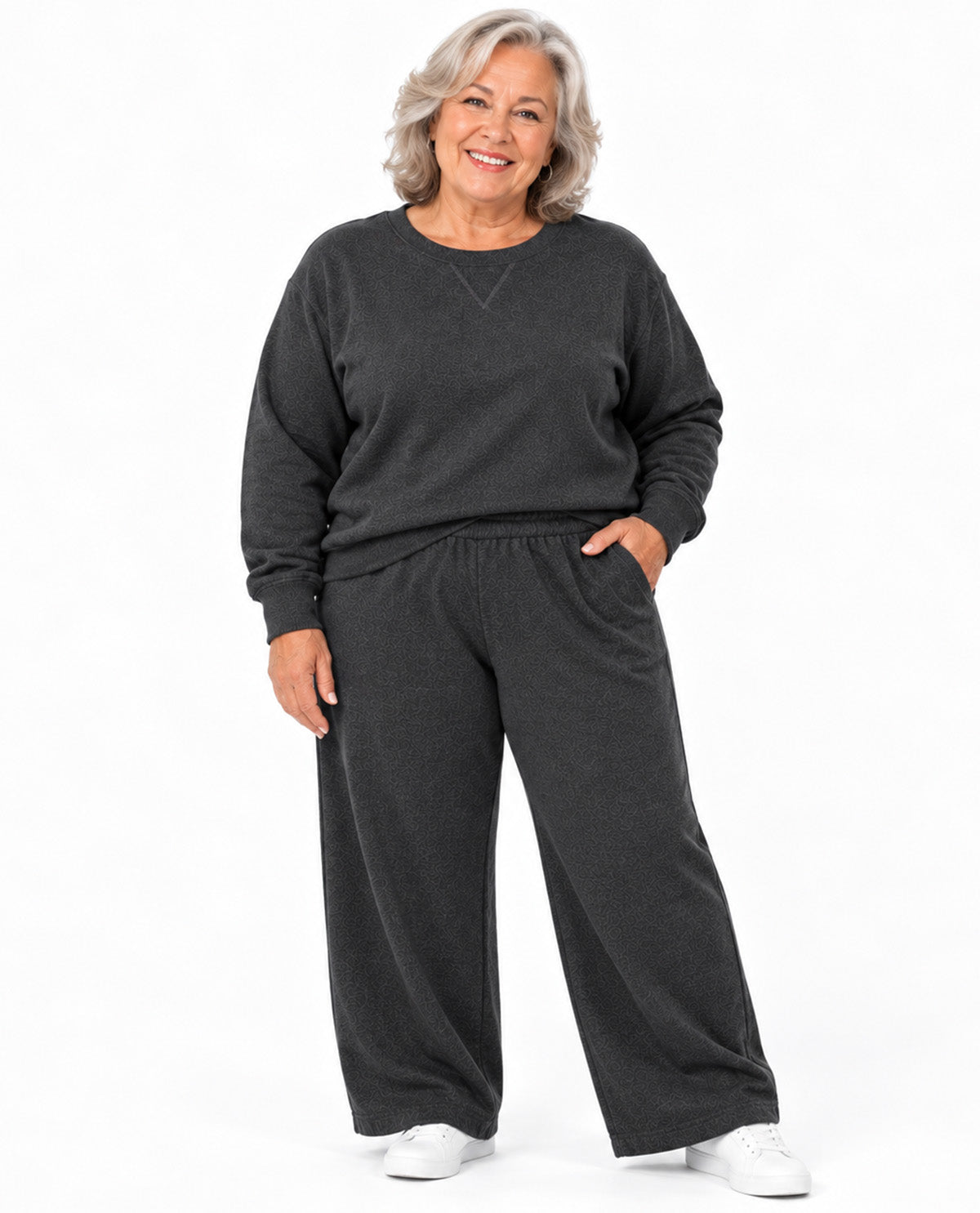 A smiling older woman stands against a plain white studio background wearing a dark charcoal gray sweatshirt and matching wide-leg sweatpants with white sneakers.