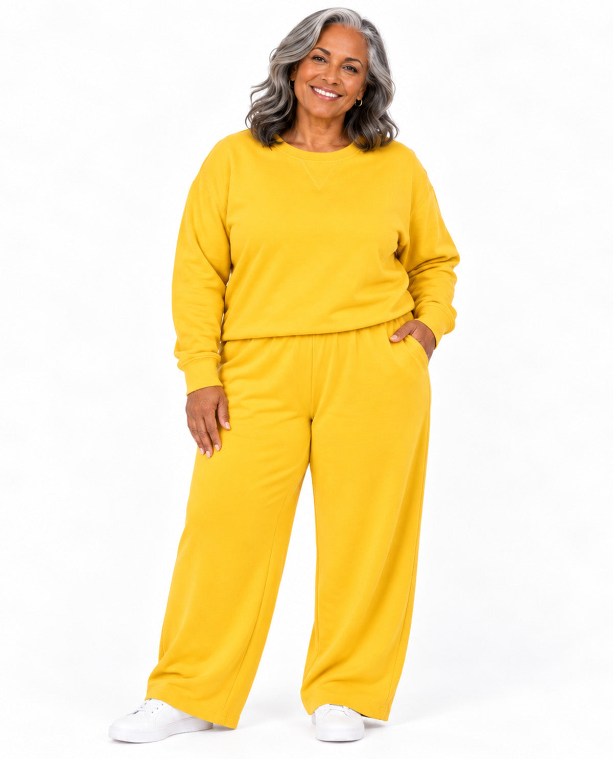 A smiling woman with gray hair wearing a matching yellow sweatshirt and wide-leg sweatpants set, paired with white sneakers, against a plain white background.