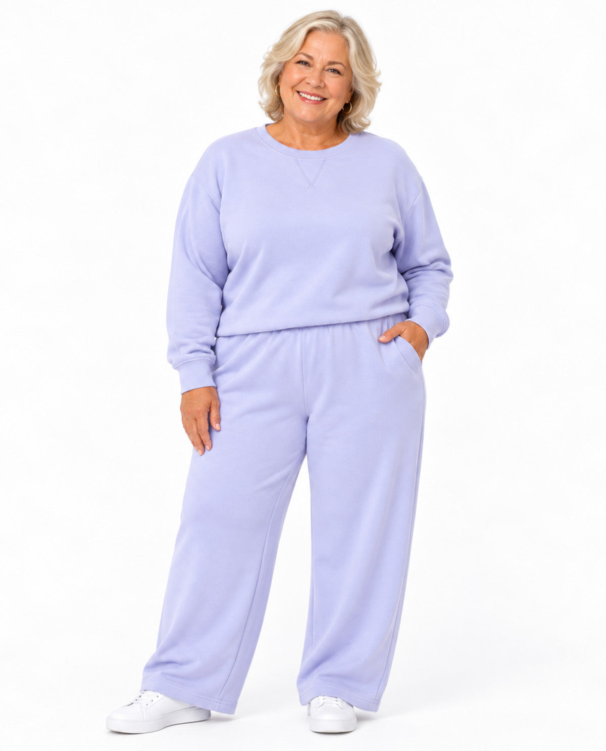 A smiling woman with blonde hair wearing a matching light lavender sweatshirt and wide-leg sweatpants set, paired with white sneakers, against a plain white studio background.
