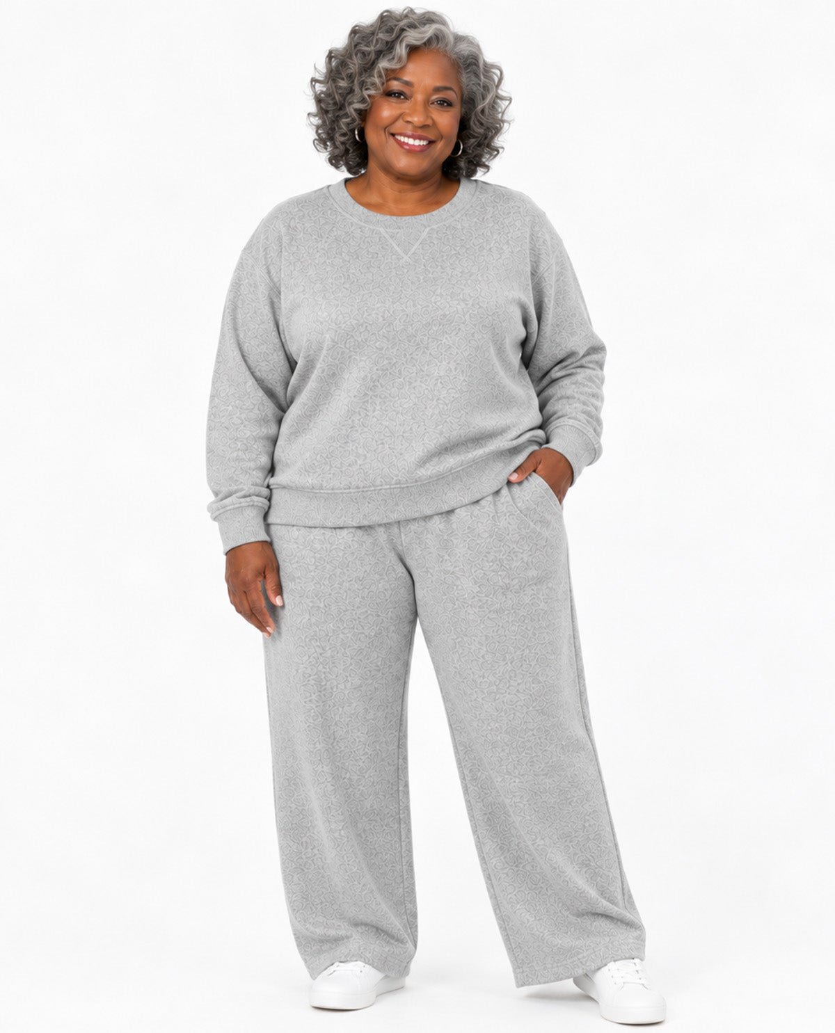 A smiling woman with curly gray hair models a matching light gray heather sweatshirt and wide-leg sweatpants set against a plain white studio background.