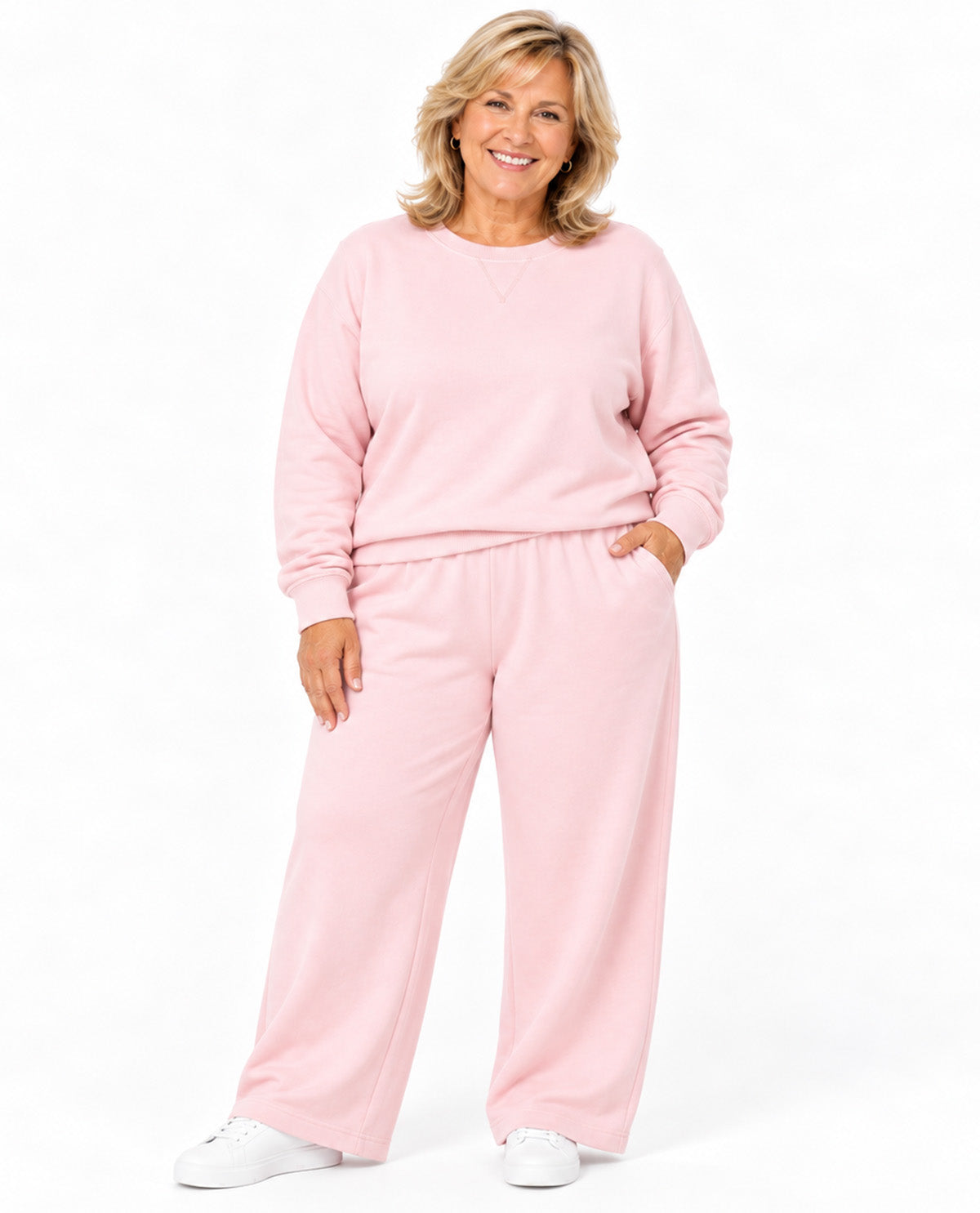 A smiling woman modeling a light pink lounge set consisting of a long-sleeve sweatshirt and matching wide-leg sweatpants, paired with white sneakers, against a plain white background.