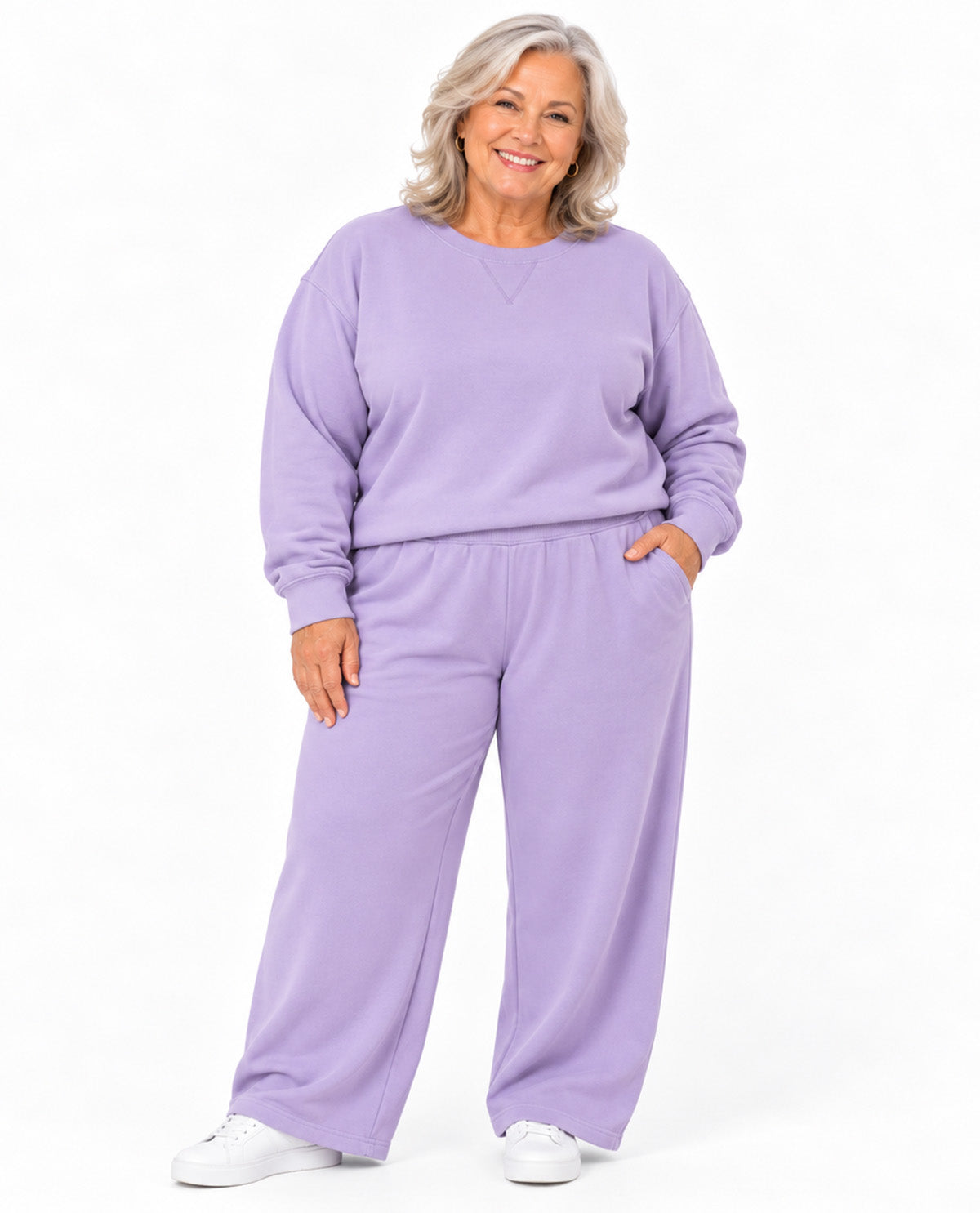 A smiling woman with gray hair models a matching lavender sweatshirt and wide-leg sweatpants set against a plain white studio background.