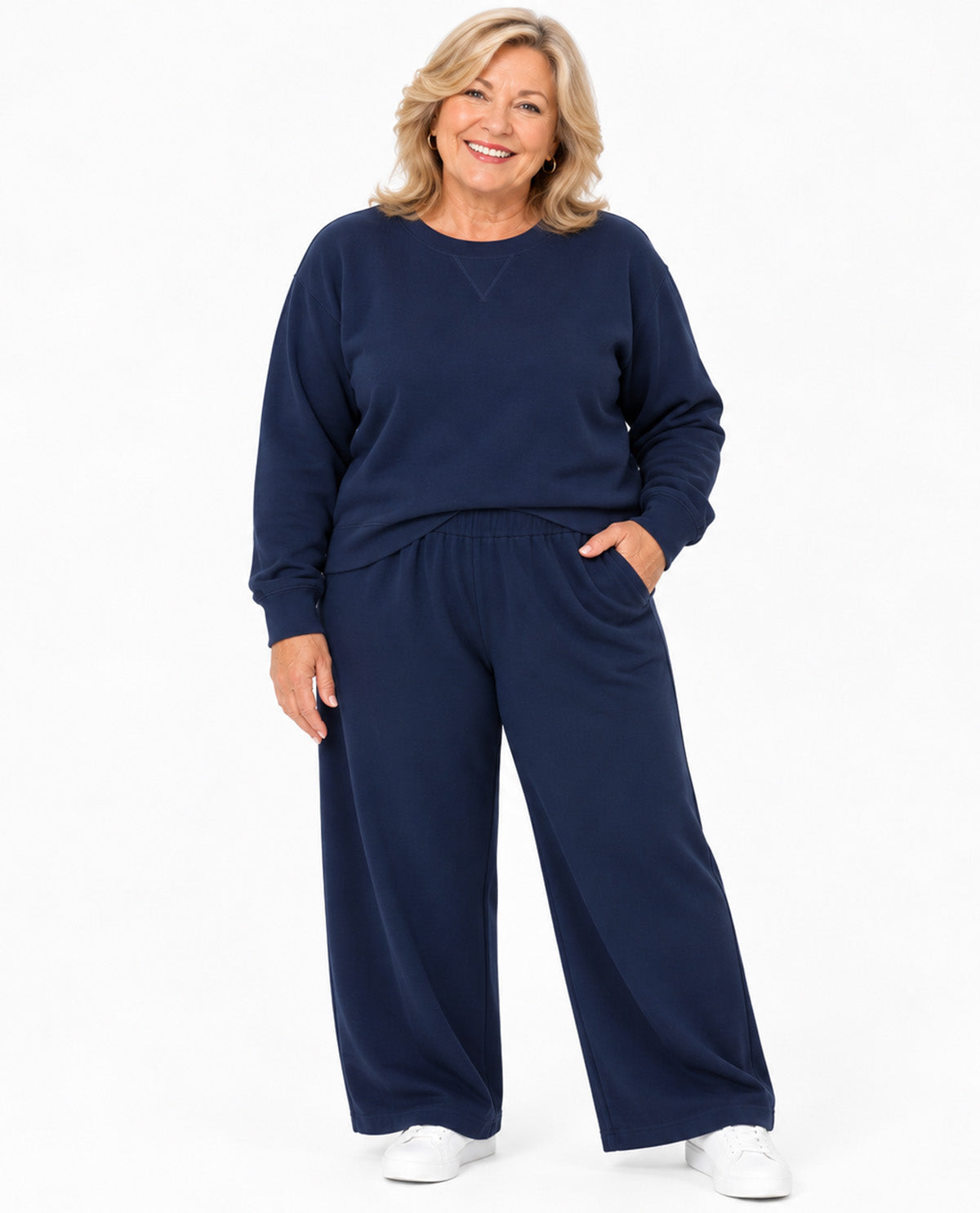 A woman modeling a matching navy blue lounge set consisting of a long-sleeve sweatshirt and wide-leg pants, paired with white sneakers, against a plain white studio background.
