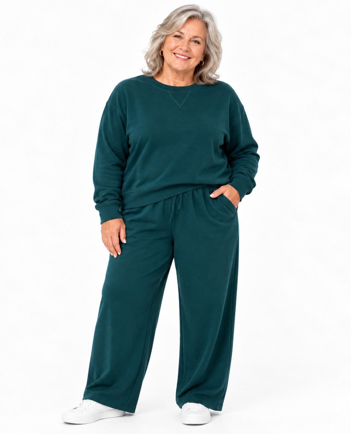 A smiling woman with gray hair stands against a plain white background wearing a dark teal green sweatshirt and matching wide-leg sweatpants, paired with white sneakers.
