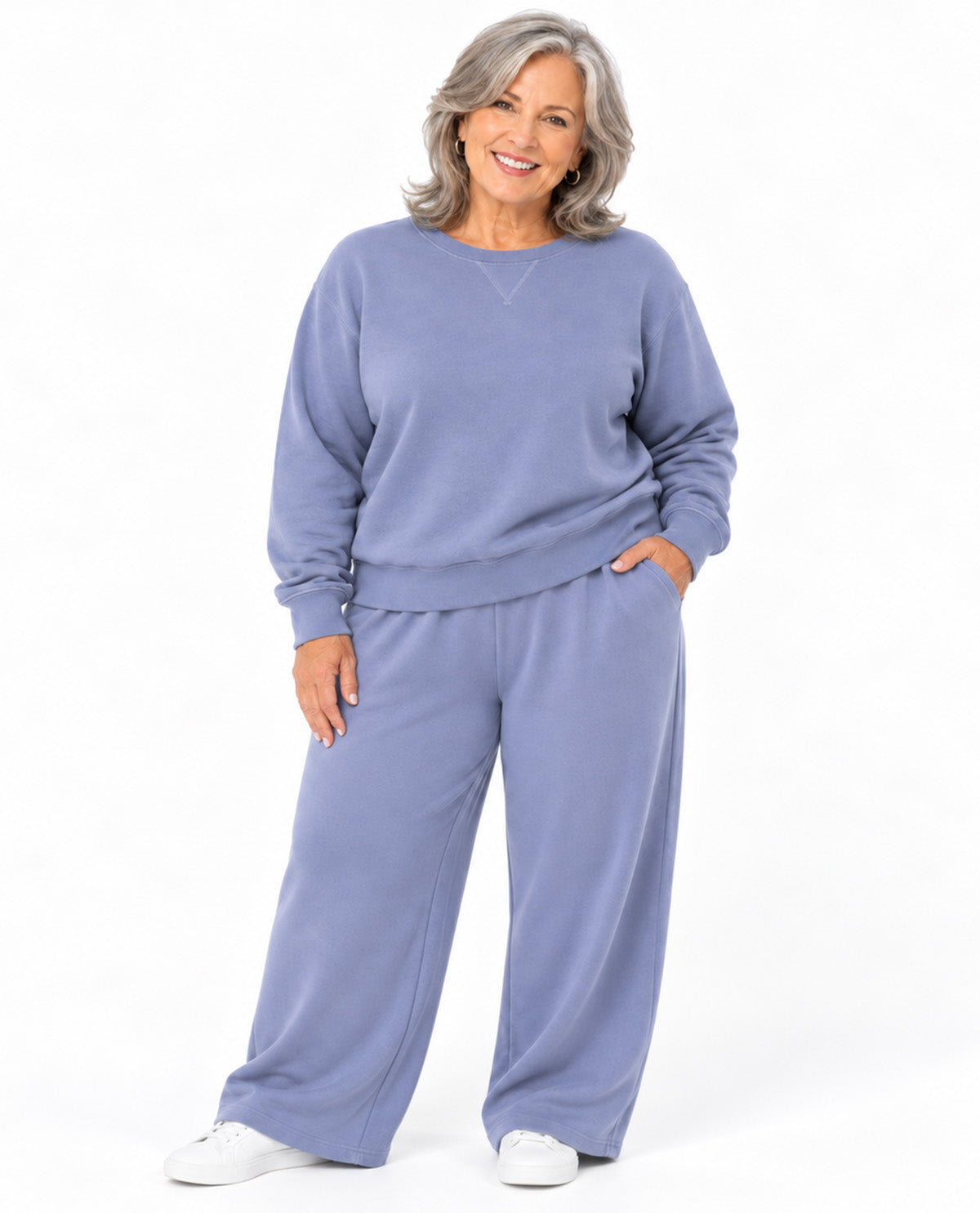 A smiling woman with gray hair models a matching two-piece lounge set consisting of a long-sleeve sweatshirt and wide-leg sweatpants in a dusty periwinkle blue color, paired with white sneakers against a white studio background.