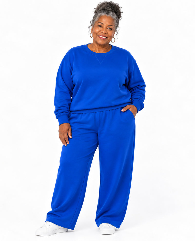 A woman modeling a matching royal blue sweatshirt and wide-leg sweatpants set, paired with white sneakers, against a plain white studio background.