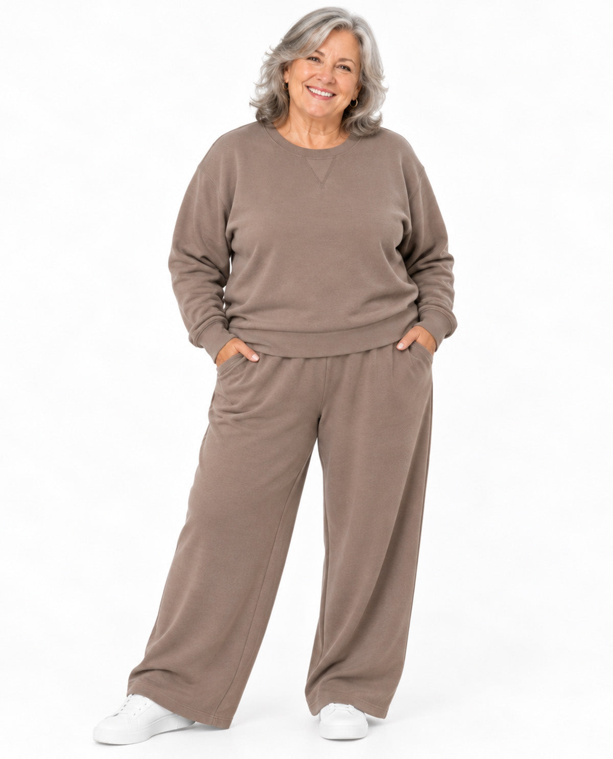 A smiling older woman wearing a matching taupe-brown lounge set consisting of a long-sleeve sweatshirt and wide-leg pants, paired with white sneakers, against a plain white background.