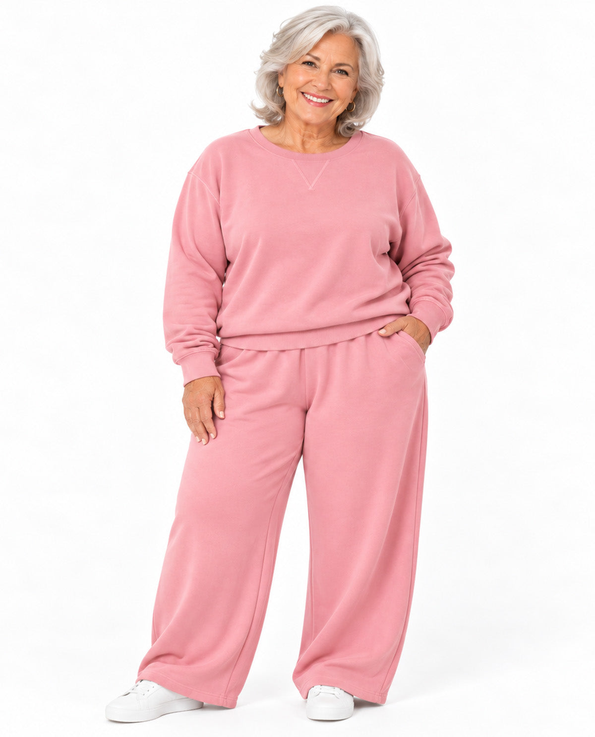 A smiling older woman with short gray hair models a matching dusty pink lounge set consisting of a long-sleeve crewneck sweatshirt and wide-leg sweatpants, paired with white sneakers against a plain white background.