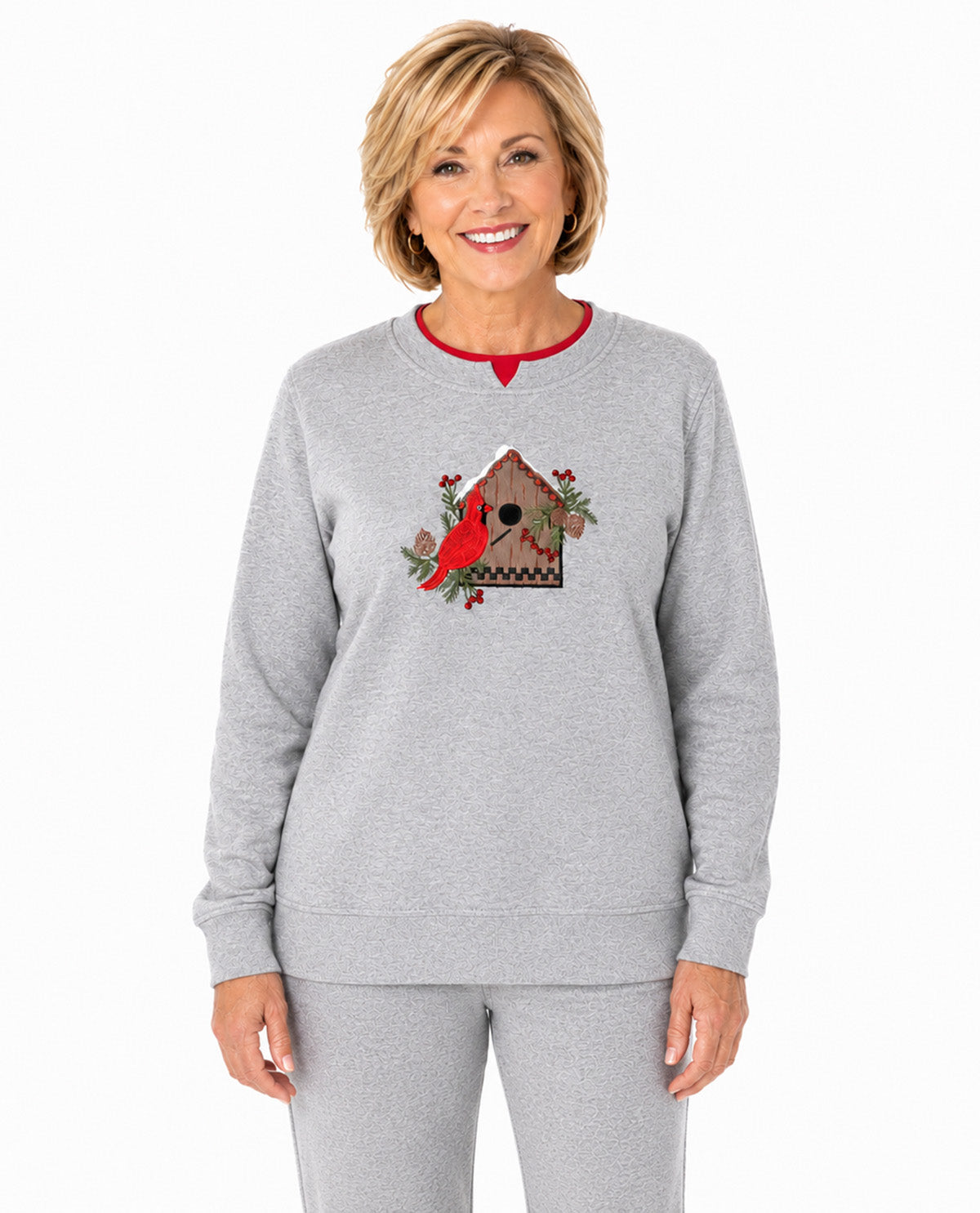 A smiling woman wearing a light heather gray crewneck sweatshirt with a red collar trim and a Christmas birdhouse graphic on the chest, paired with matching gray sweatpants, against a plain white background.