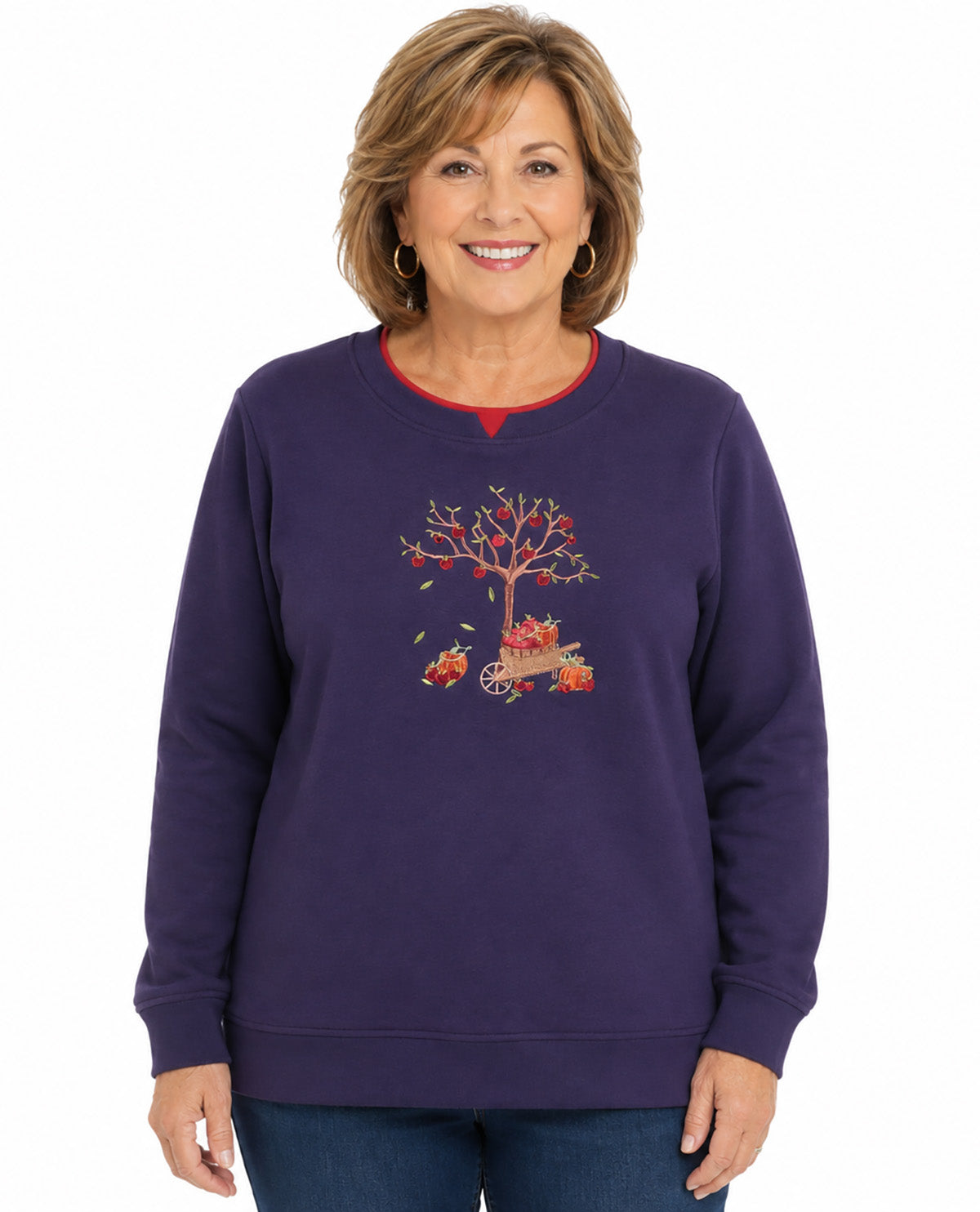 A woman wearing a purple crewneck sweatshirt with a red inner collar and a graphic of a tree with red leaves and a small red car on the front, paired with blue jeans, against a plain white background.