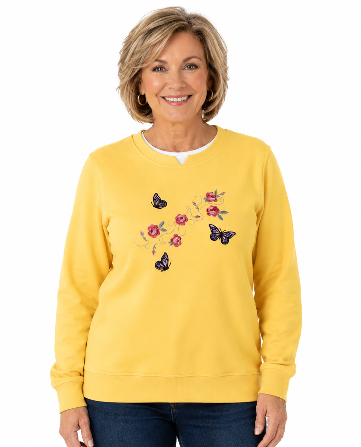 A woman wearing a bright yellow crewneck sweatshirt with embroidered pink flowers and black butterflies on the front, paired with dark blue jeans, against a plain white background.