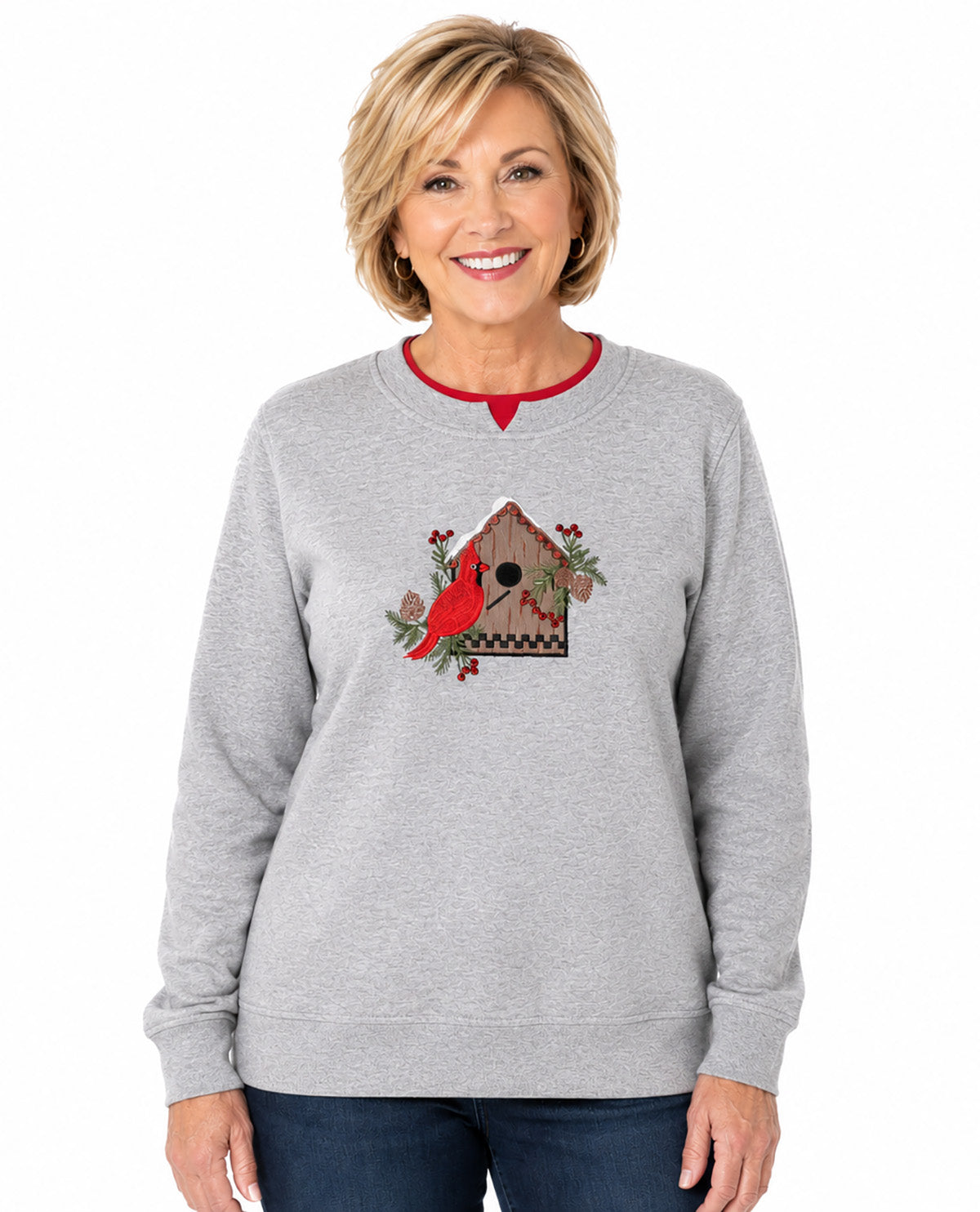 A smiling blonde woman wearing a light heather gray crewneck sweatshirt with a red collar trim and a central graphic of a red cardinal perched on a birdhouse with greenery, paired with dark blue jeans against a plain white background.