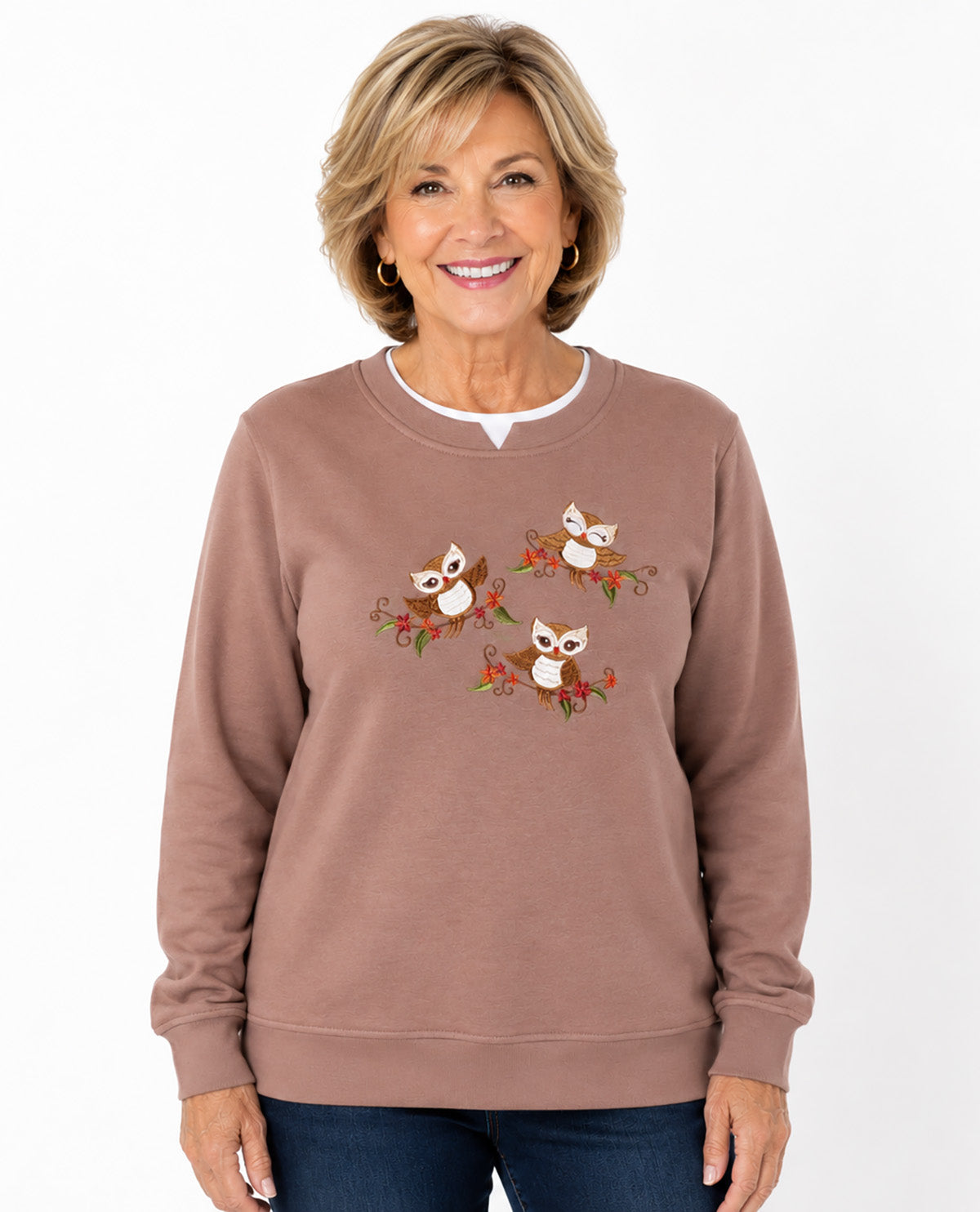 A smiling woman with short blonde hair wearing a dusty rose crewneck sweatshirt with a graphic of three cartoon owls on branches, paired with dark blue jeans, against a plain white background.