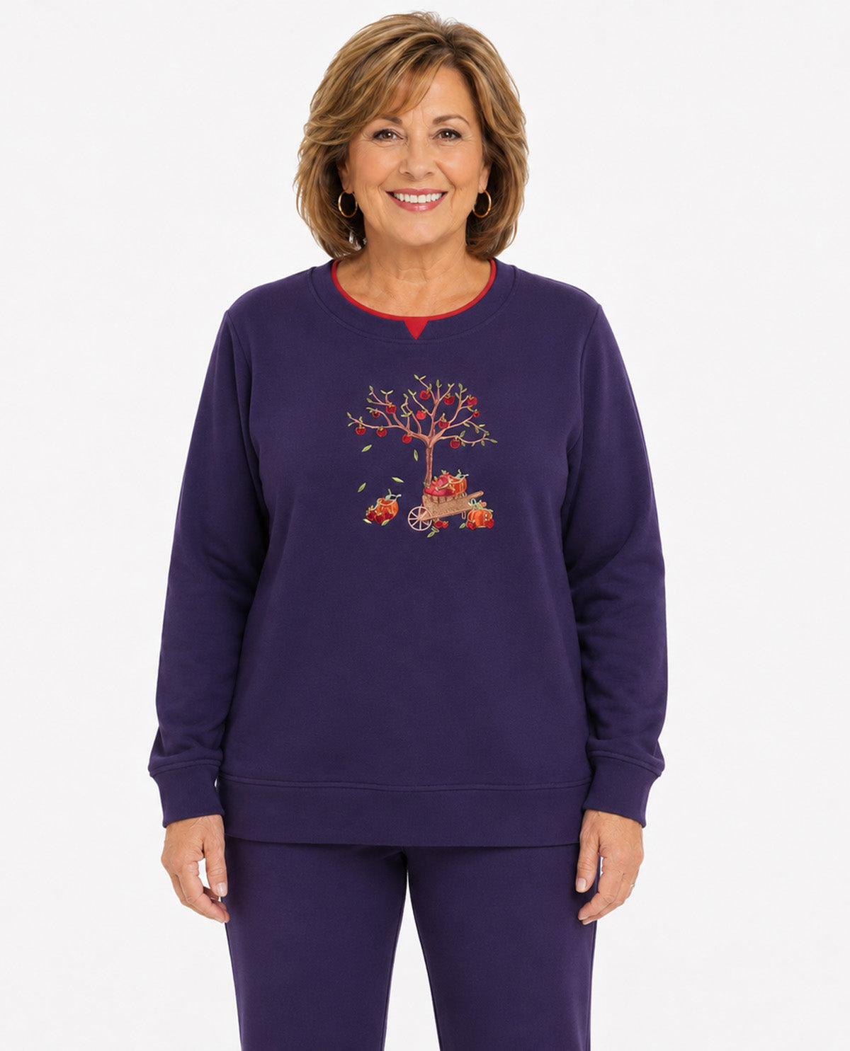 A smiling woman wearing a dark purple crewneck sweatshirt and matching sweatpants. The sweatshirt features a colorful graphic of an autumn tree with red leaves and a small red vehicle on the front, with a thin red inner collar.