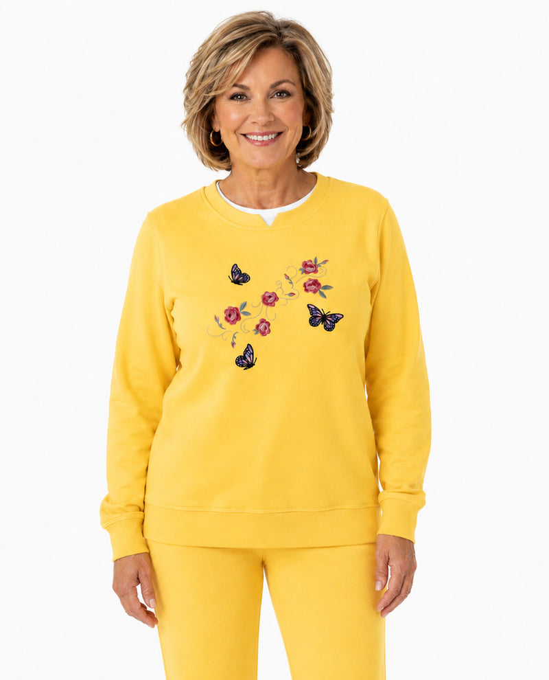 A smiling woman wearing a bright yellow crewneck sweatshirt with embroidered butterflies and flowers on the chest, paired with matching yellow pants, against a plain white background.