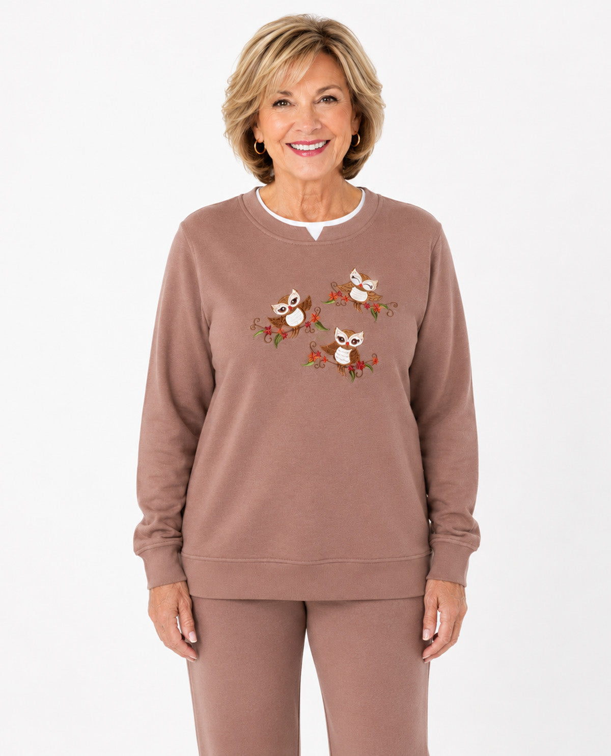 A smiling woman wearing a dusty rose crewneck sweatshirt with three embroidered owl motifs on the chest, paired with matching sweatpants, against a plain white background.