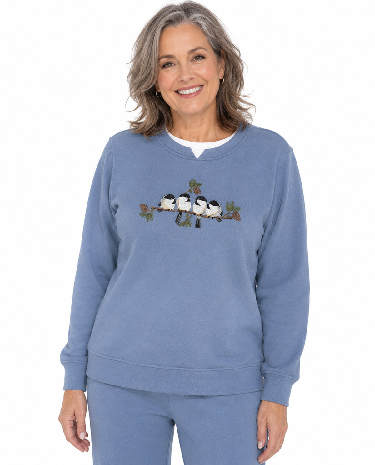 A smiling woman wearing a dusty blue crewneck sweatshirt with a graphic of four birds on a branch across the chest, paired with matching blue sweatpants, against a plain white background.