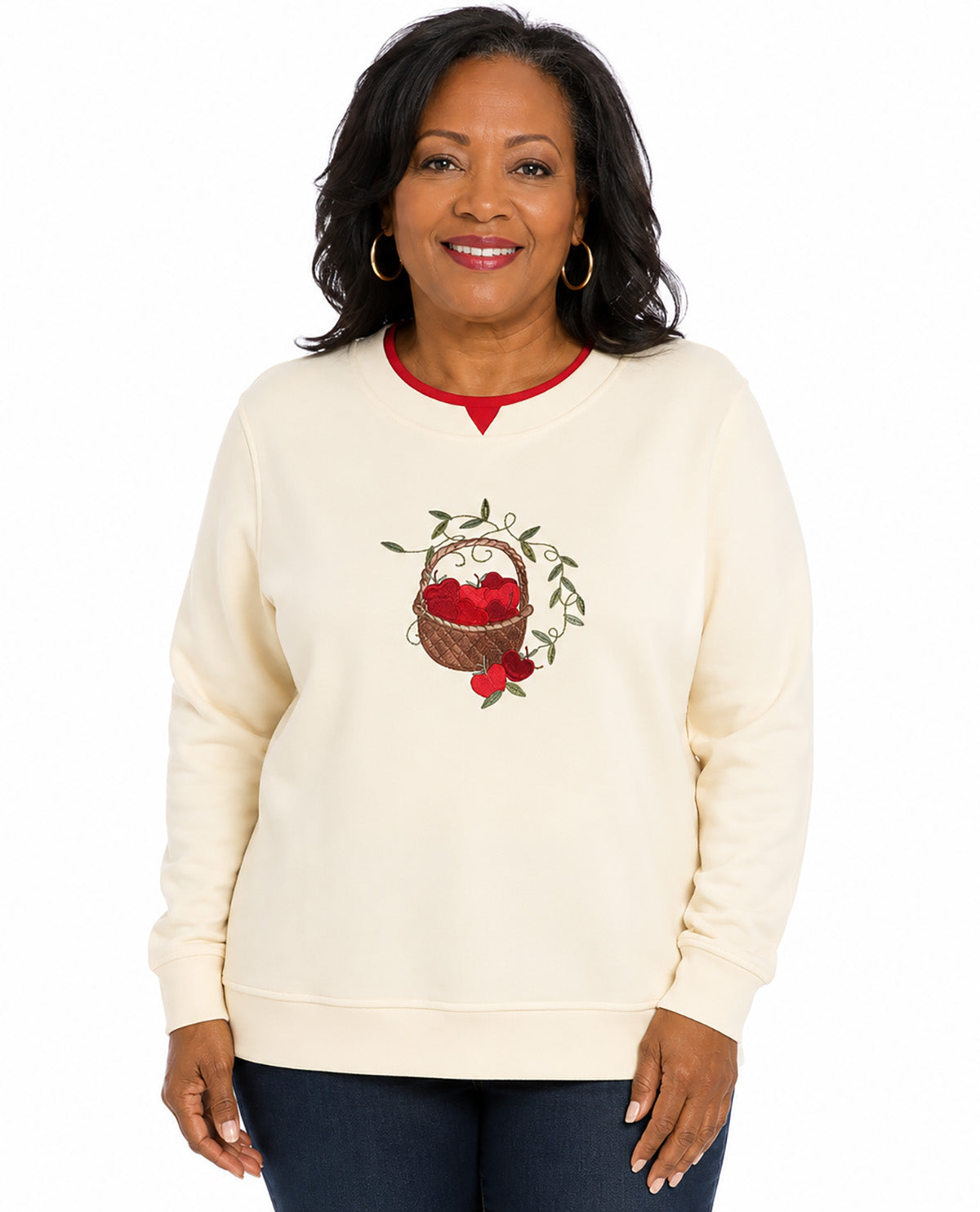 A woman wearing a cream-colored crewneck sweatshirt with a red collar trim and a central graphic of a basket of red apples with green leaves. She is also wearing dark blue jeans and gold hoop earrings, photographed against a solid white background.