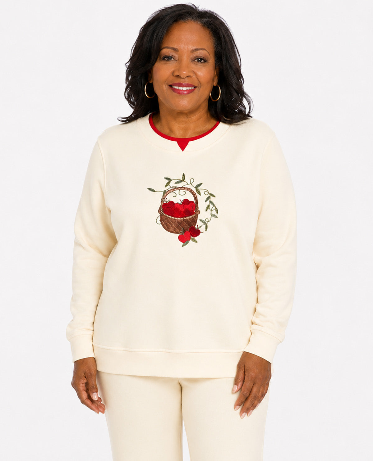 A woman modeling a cream-colored sweatshirt and matching sweatpants set. The sweatshirt features a red crew neck, a red berry basket graphic on the chest, and red cuffs and hem.
