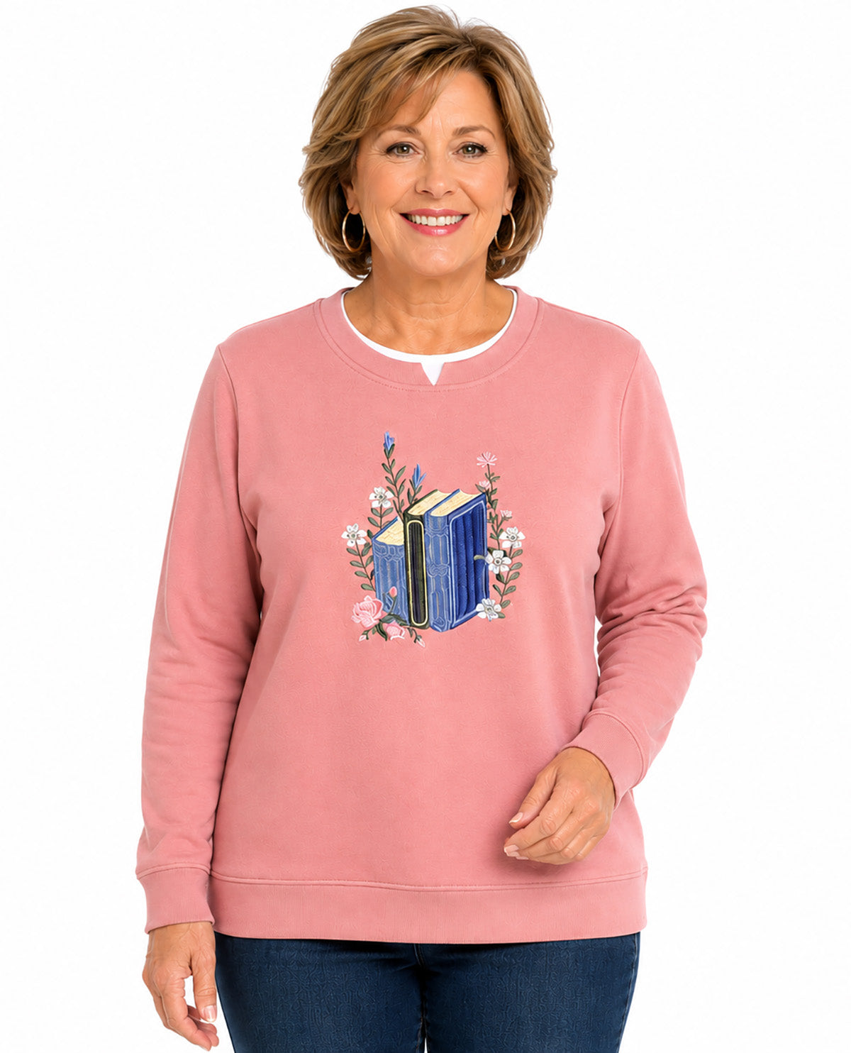 A smiling woman wearing a dusty pink crewneck sweatshirt with a graphic of a blue book surrounded by pink flowers on the chest, paired with dark blue jeans, against a plain white background.