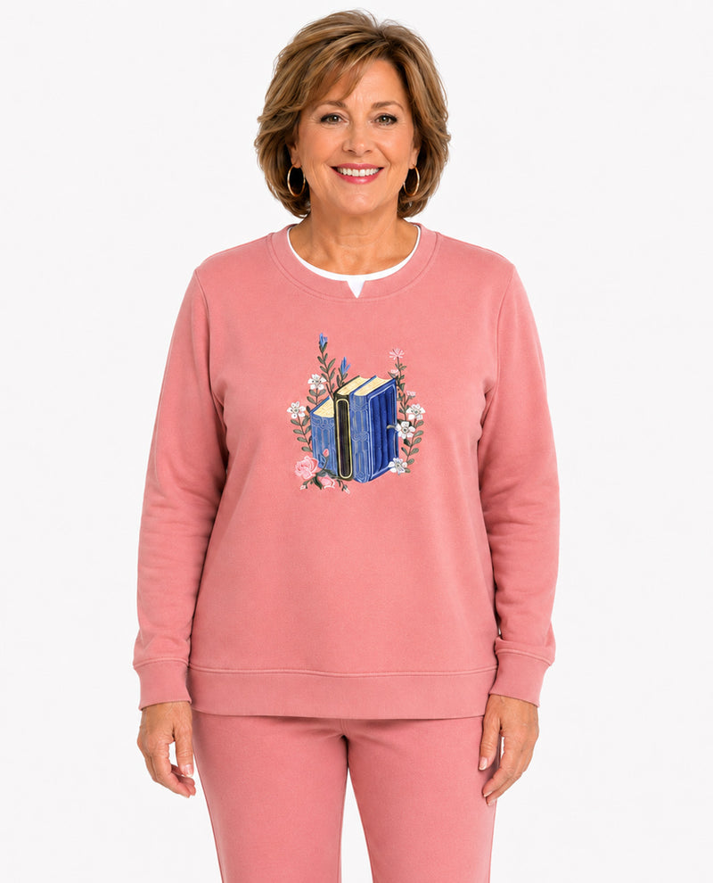 A smiling woman wearing a dusty pink crewneck sweatshirt and matching sweatpants. The sweatshirt features a central graphic of a blue book surrounded by pink flowers.