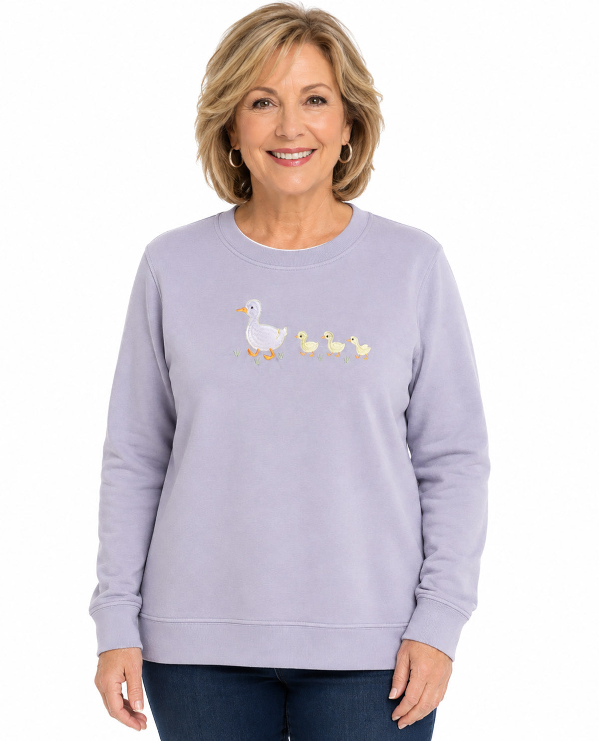 A woman wearing a light lavender crewneck sweatshirt with a graphic of a white goose and three yellow ducklings on the chest, paired with dark blue jeans, against a plain white background.