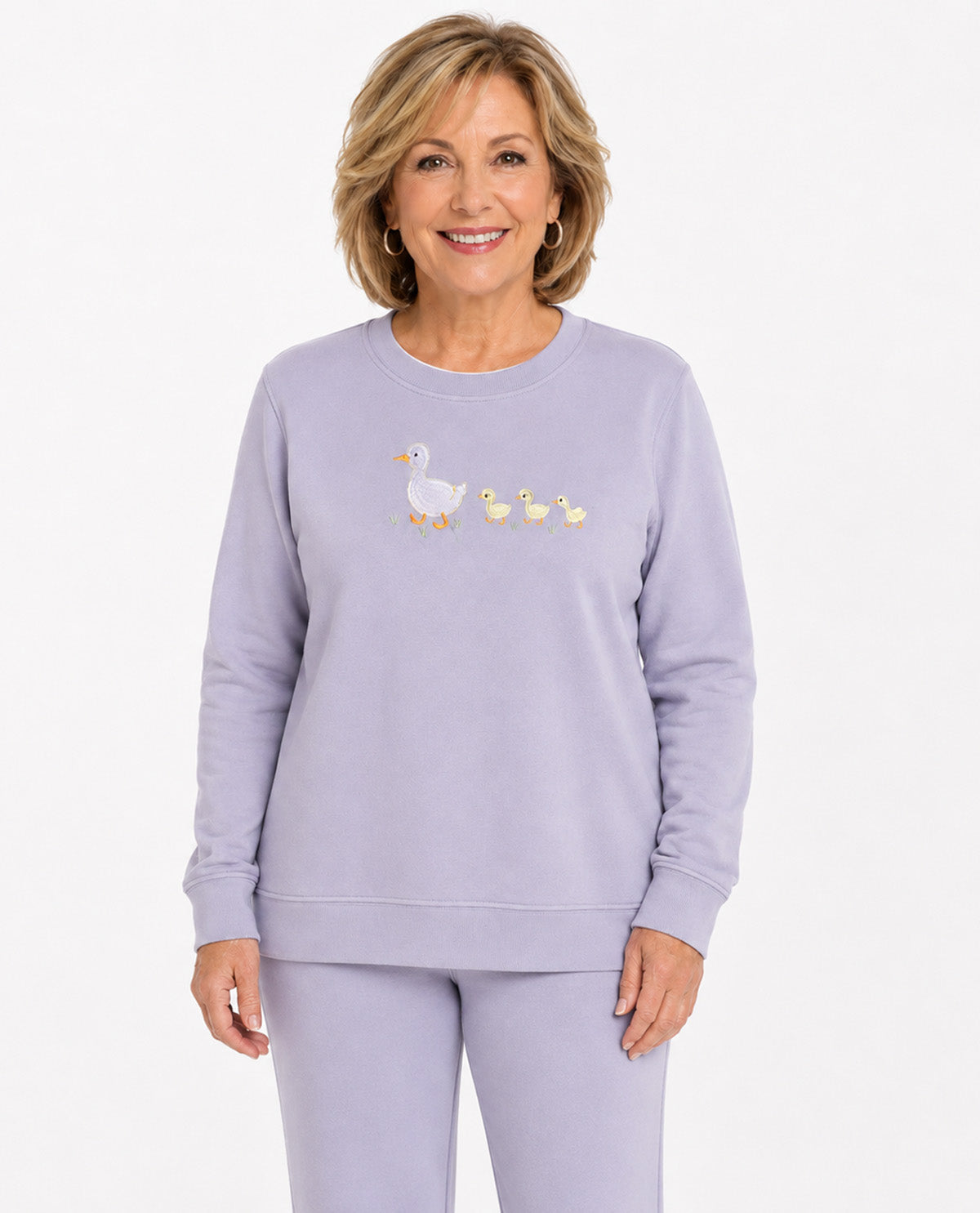 A smiling woman wearing a light lavender crewneck sweatshirt and matching sweatpants. The sweatshirt features a small embroidered graphic of a white duck with three yellow ducklings on the left chest.