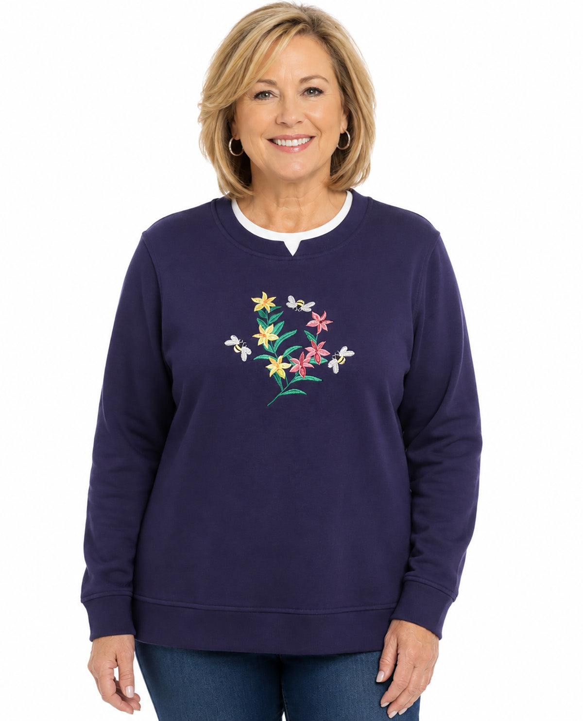 A woman wearing a navy blue crewneck sweatshirt with a colorful embroidered floral design on the chest, paired with blue jeans, against a plain white background.