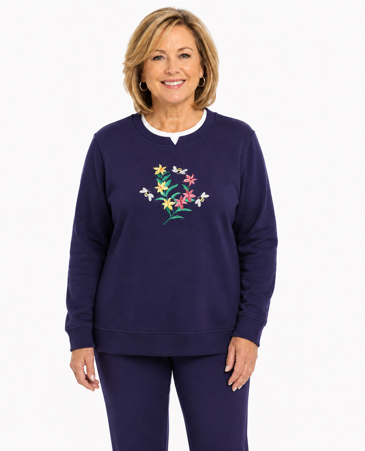 A woman modeling a navy blue sweatshirt and matching sweatpants set. The sweatshirt features a colorful embroidered floral design on the center chest, with a white inner collar.
