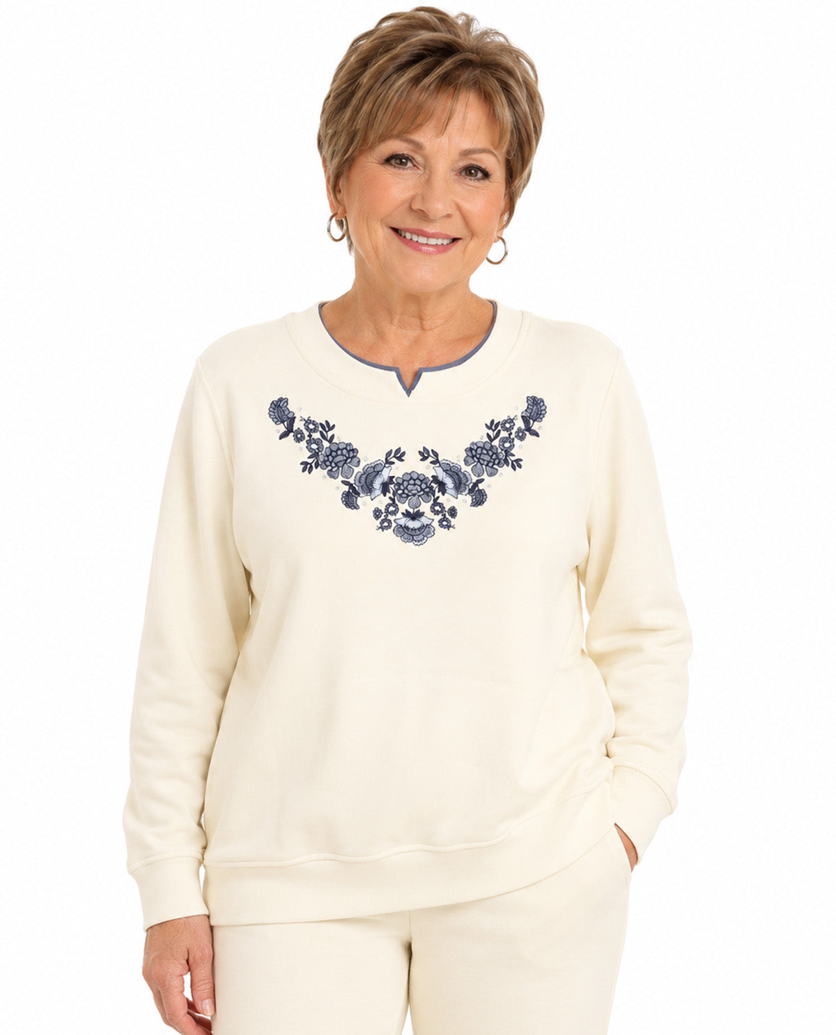 An older woman modeling a cream-colored crewneck sweatshirt with a blue floral embroidered pattern across the chest, paired with matching cream pants, against a plain white background.