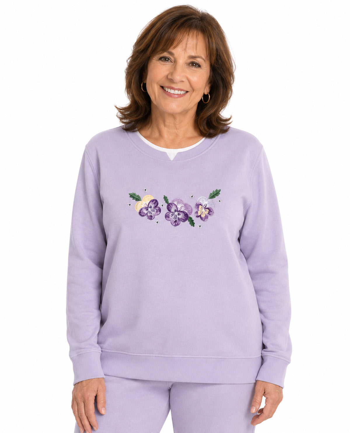 A smiling woman wearing a light lavender crewneck sweatshirt with a colorful embroidered pansy design on the chest, paired with matching lavender sweatpants, against a plain white background.