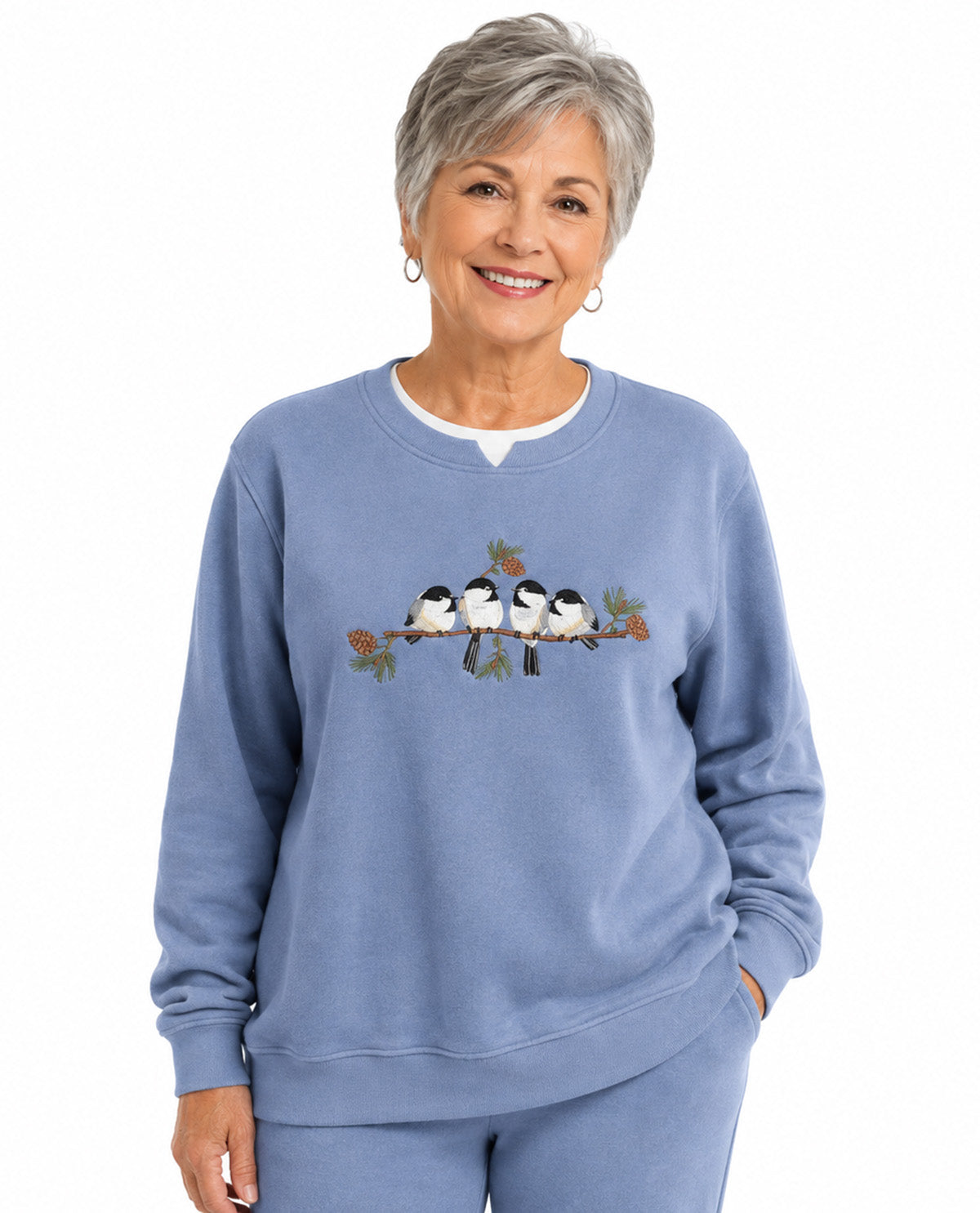 An older woman with short gray hair smiling while wearing a light blue crewneck sweatshirt with a graphic of four birds on a branch across the chest, paired with matching light blue sweatpants against a white background.