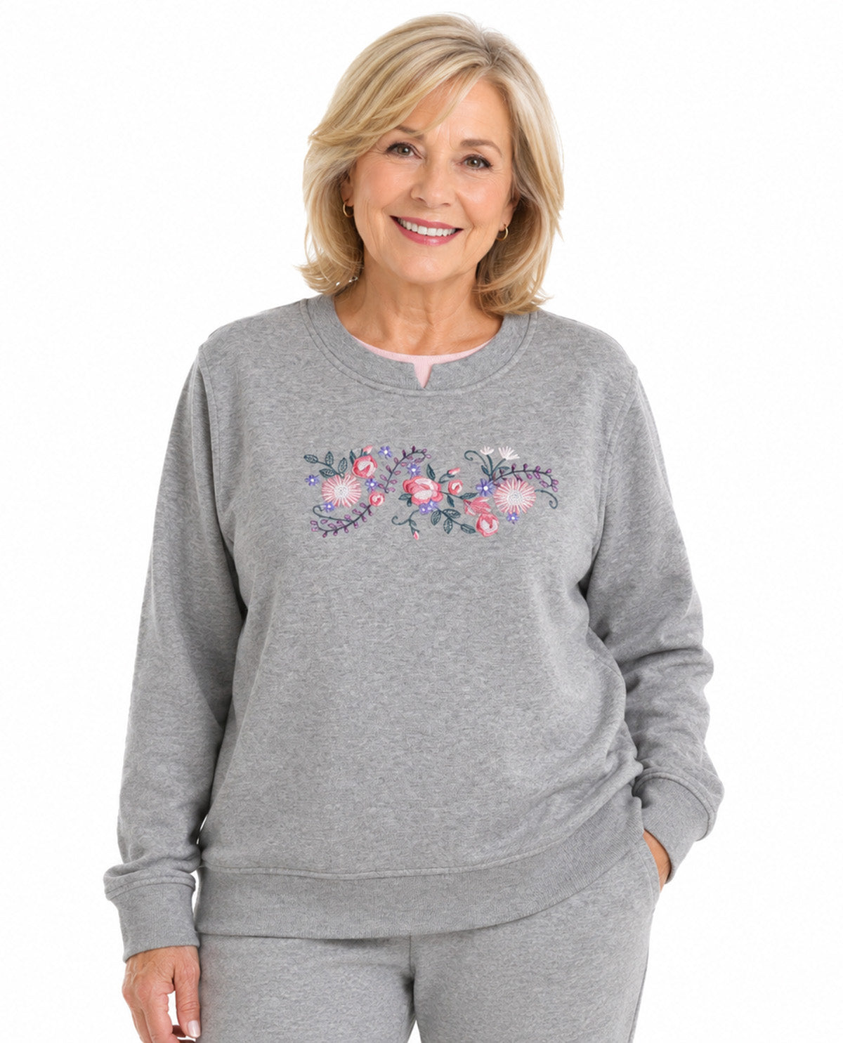 A smiling blonde woman wearing a heather gray crewneck sweatshirt with a colorful floral and bird pattern across the chest, paired with matching gray sweatpants, against a plain white background.