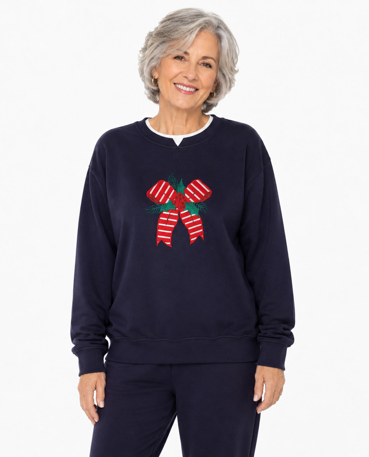 A smiling older woman with short gray hair wears a navy crewneck sweatshirt featuring a red and white striped candy cane bow graphic on the chest, paired with matching navy pants, against a plain white background.
