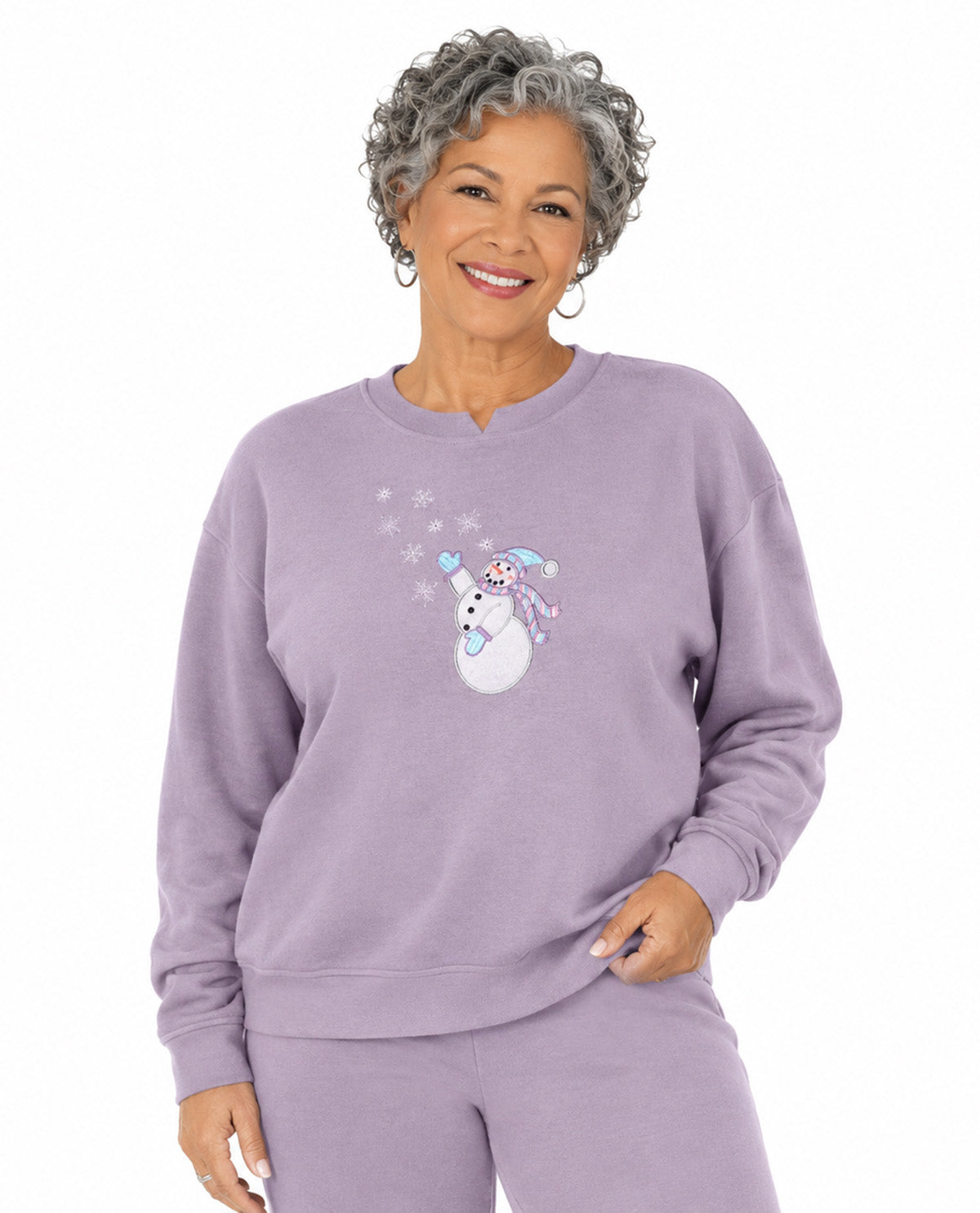 A smiling woman with short gray curly hair wears a light lavender crewneck sweatshirt and matching sweatpants. The sweatshirt features a graphic of a snowman wearing a blue hat and scarf, surrounded by small white snowflakes.