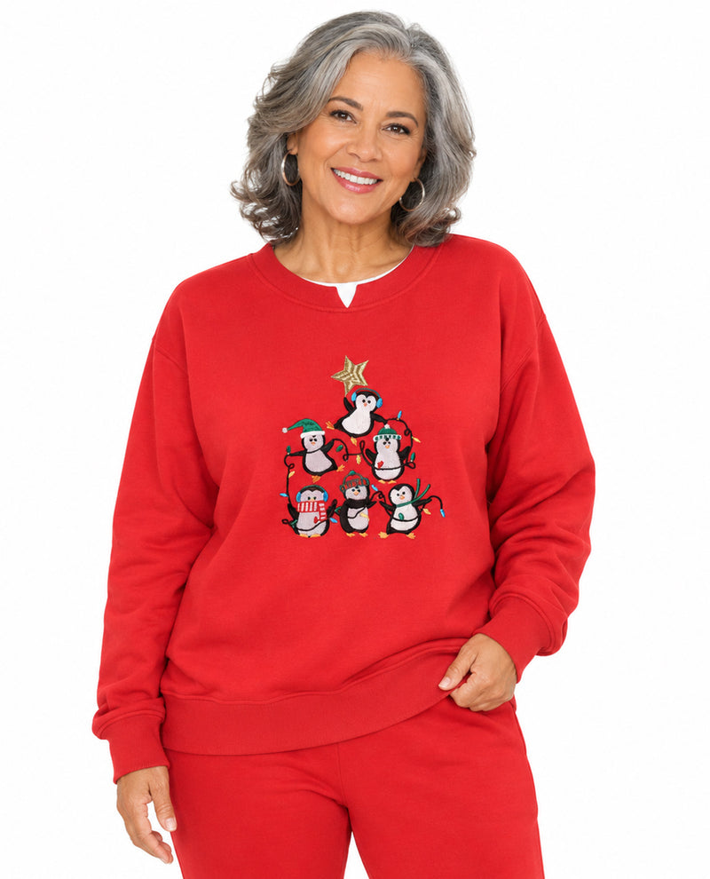 A smiling woman with gray hair wearing a bright red crewneck sweatshirt and matching red sweatpants. The sweatshirt features a graphic of cartoon penguins dressed in Christmas attire under a star.