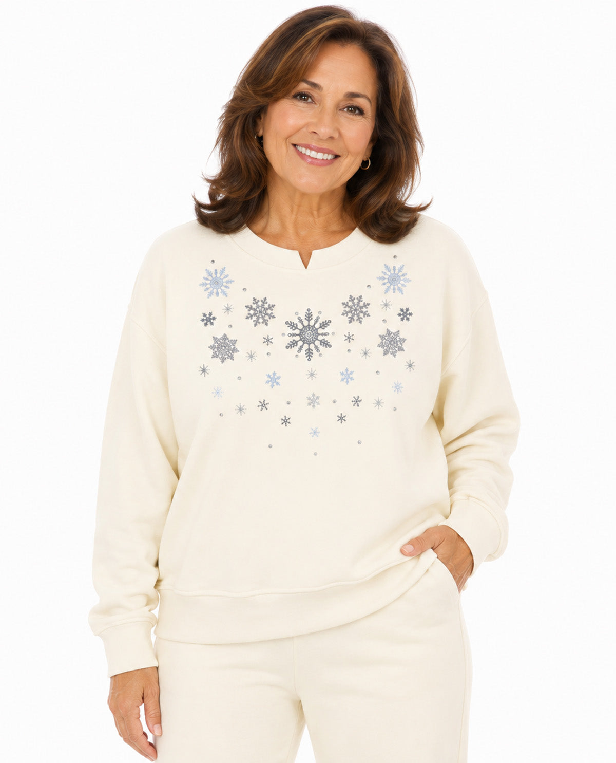 A smiling woman wearing a cream-colored crewneck sweatshirt with a festive snowflake pattern in light blue and gray on the front, paired with matching cream sweatpants, set against a plain white background.