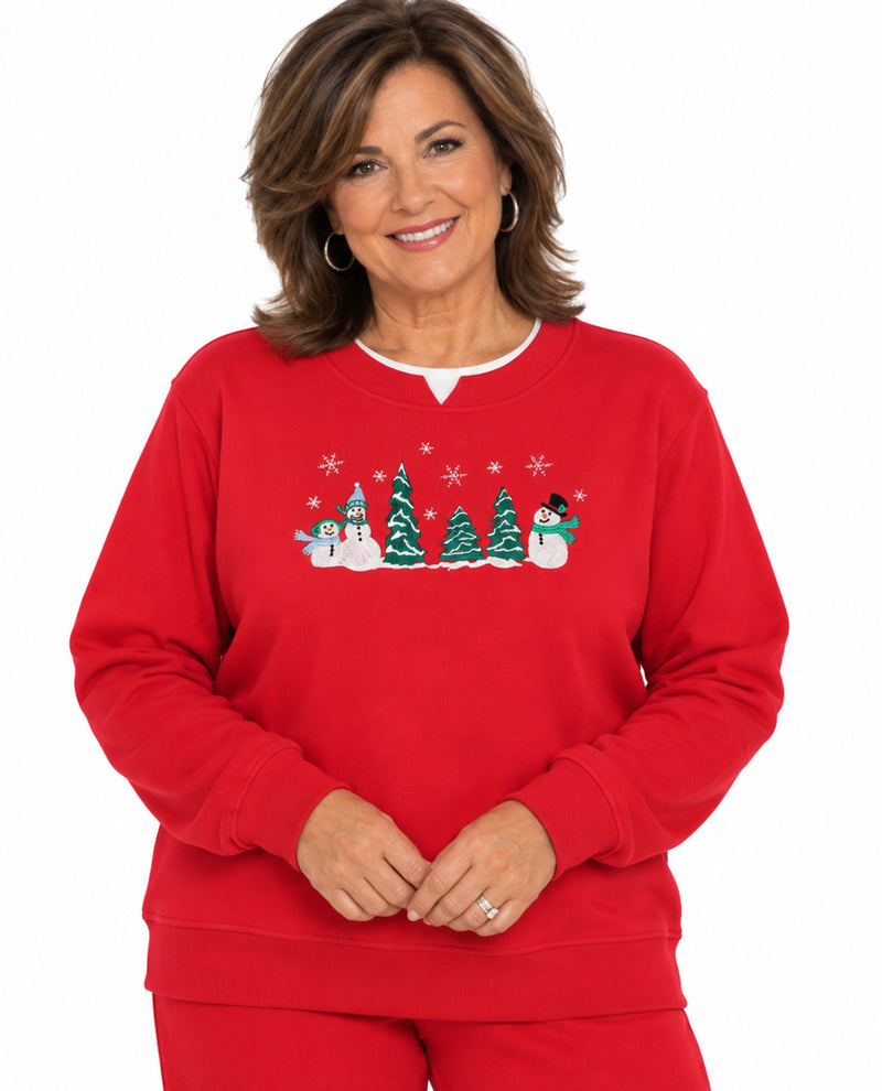 A smiling woman wearing a bright red crewneck sweatshirt with a festive Christmas tree and snowman graphic across the chest, paired with matching red pants, against a plain white background.