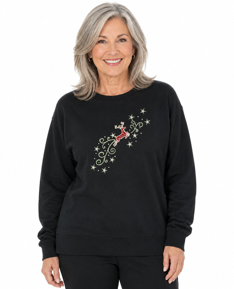 A smiling woman with shoulder-length gray hair wears a black crewneck sweatshirt featuring a diagonal graphic of a red reindeer, white stars, and gold swirls on the chest, set against a plain white background.