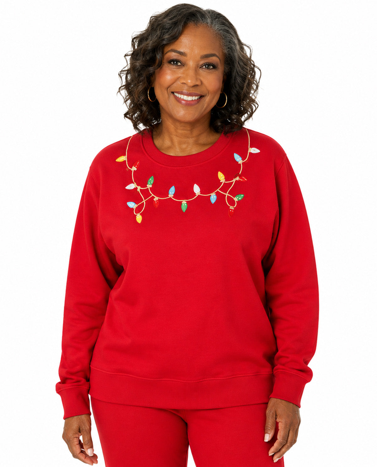 A smiling woman wearing a bright red crewneck sweatshirt with a colorful Christmas light garland embroidered design across the chest, paired with matching red pants, against a plain white background.