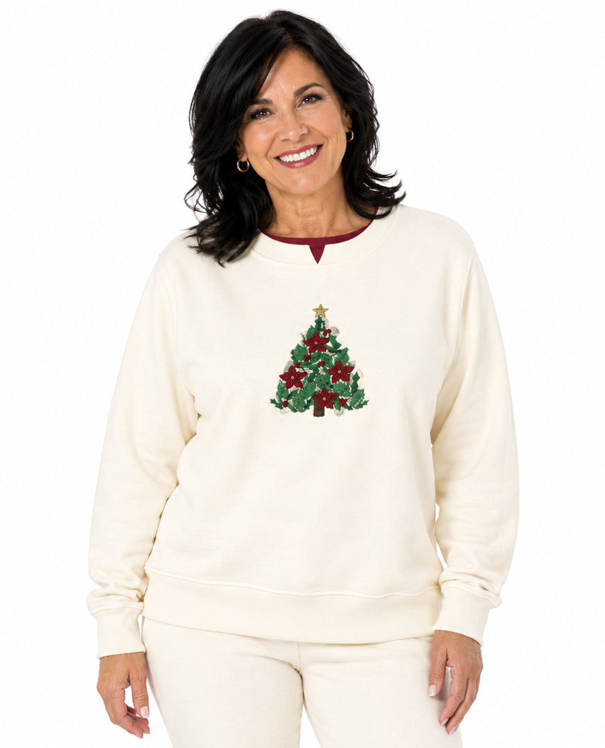A smiling woman wearing a cream-colored crewneck sweatshirt with a green and red Christmas tree graphic on the chest, paired with matching cream pants, set against a plain white background.