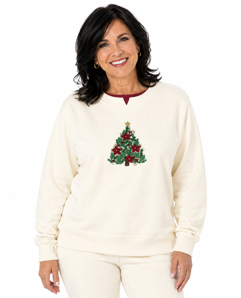 A smiling woman wearing a cream-colored crewneck sweatshirt with a green and red Christmas tree graphic on the chest, paired with matching cream pants, set against a plain white background.