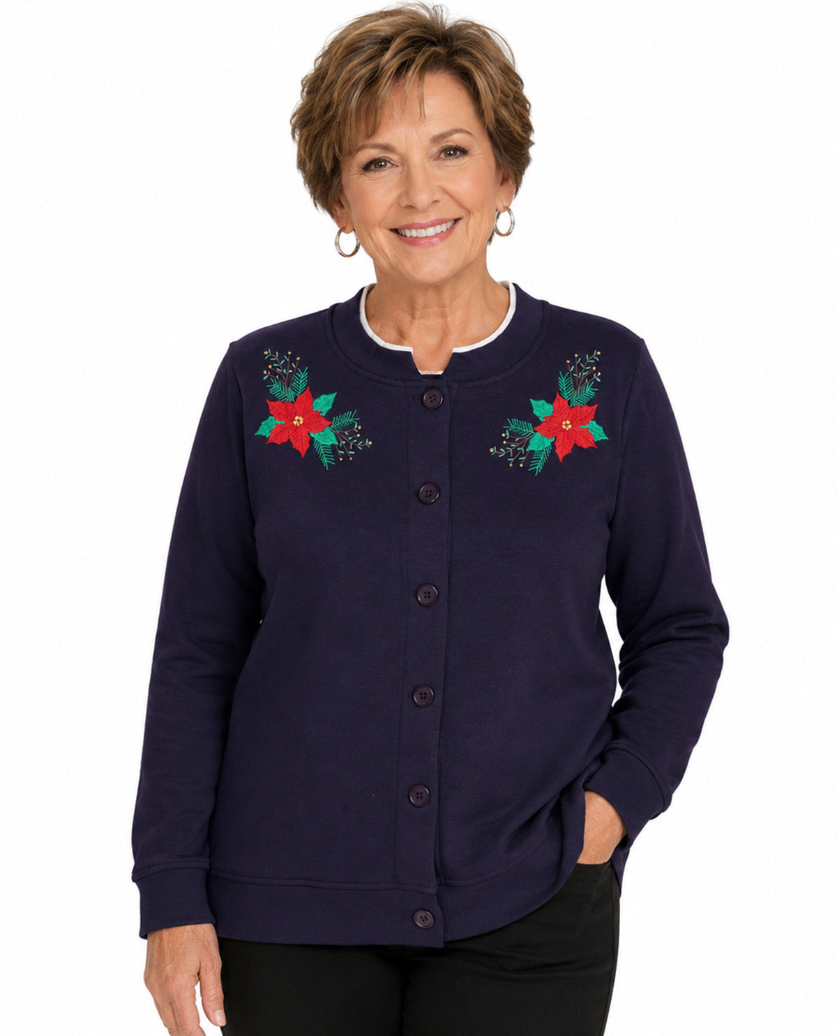 A smiling older woman wearing a navy blue button-front cardigan with embroidered red poinsettias and green leaves on the chest, paired with black pants, against a plain white background.