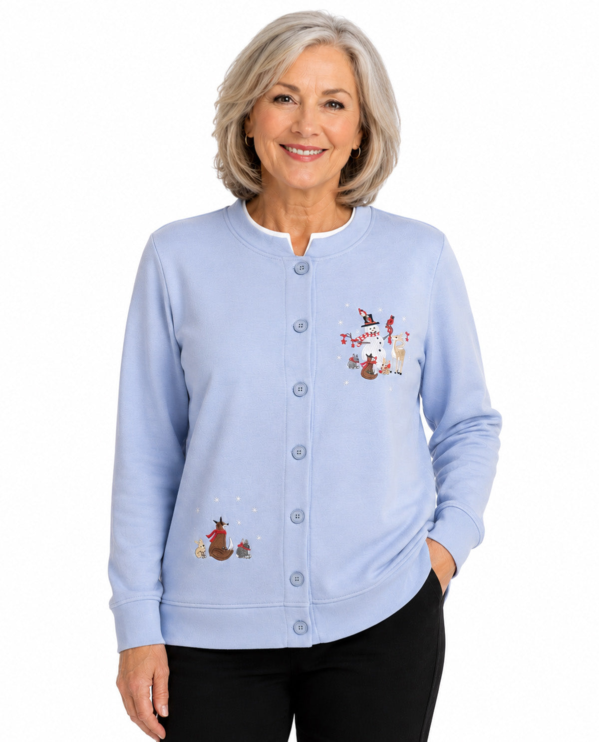 A smiling older woman with short gray hair wears a light blue button-front cardigan with small illustrated woodland character prints on the chest, paired with black pants, against a plain white studio background.
