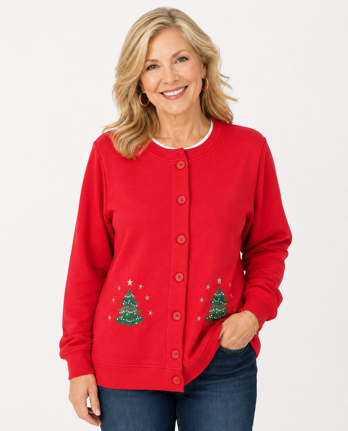 A smiling woman wearing a bright red button-front cardigan sweater with embroidered green Christmas trees and small white stars on the lower front, paired with dark blue jeans, against a plain light gray studio background.
