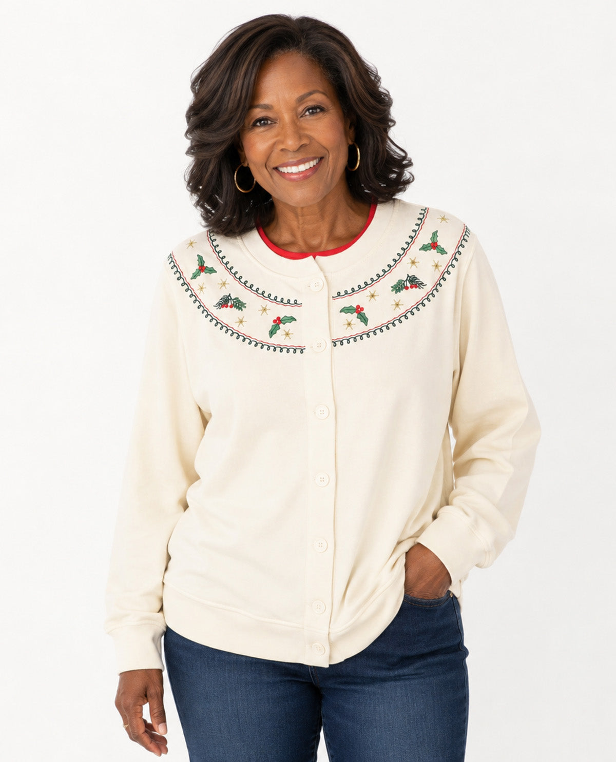 A smiling woman wearing a cream-colored button-up cardigan with embroidered holiday motifs around the collar, paired with dark blue jeans, set against a plain white studio background.
