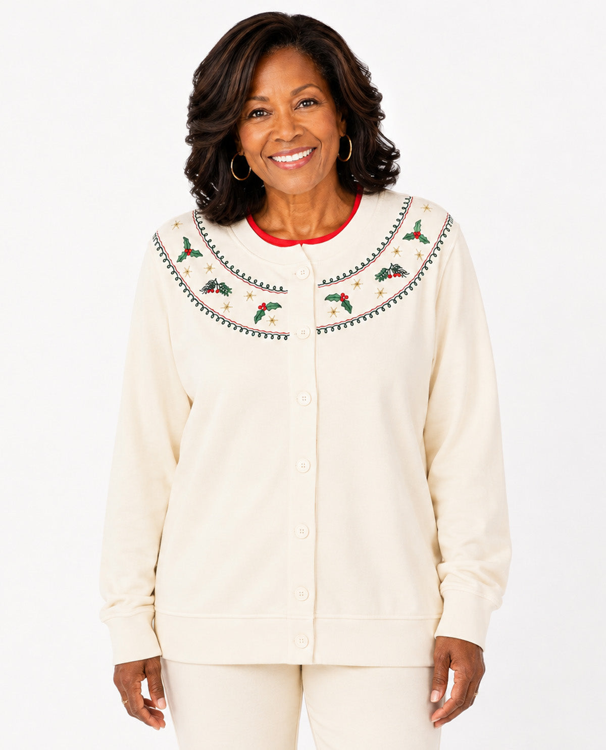 A smiling woman wearing a cream-colored button-up cardigan with a large decorative yoke featuring embroidered holly leaves, pinecones, and red berries, paired with matching cream pants against a white studio background.