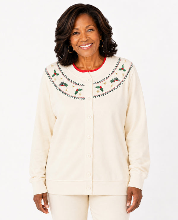 A smiling woman wearing a cream-colored button-up cardigan with a large decorative yoke featuring embroidered holly leaves, pinecones, and red berries, paired with matching cream pants against a white studio background.