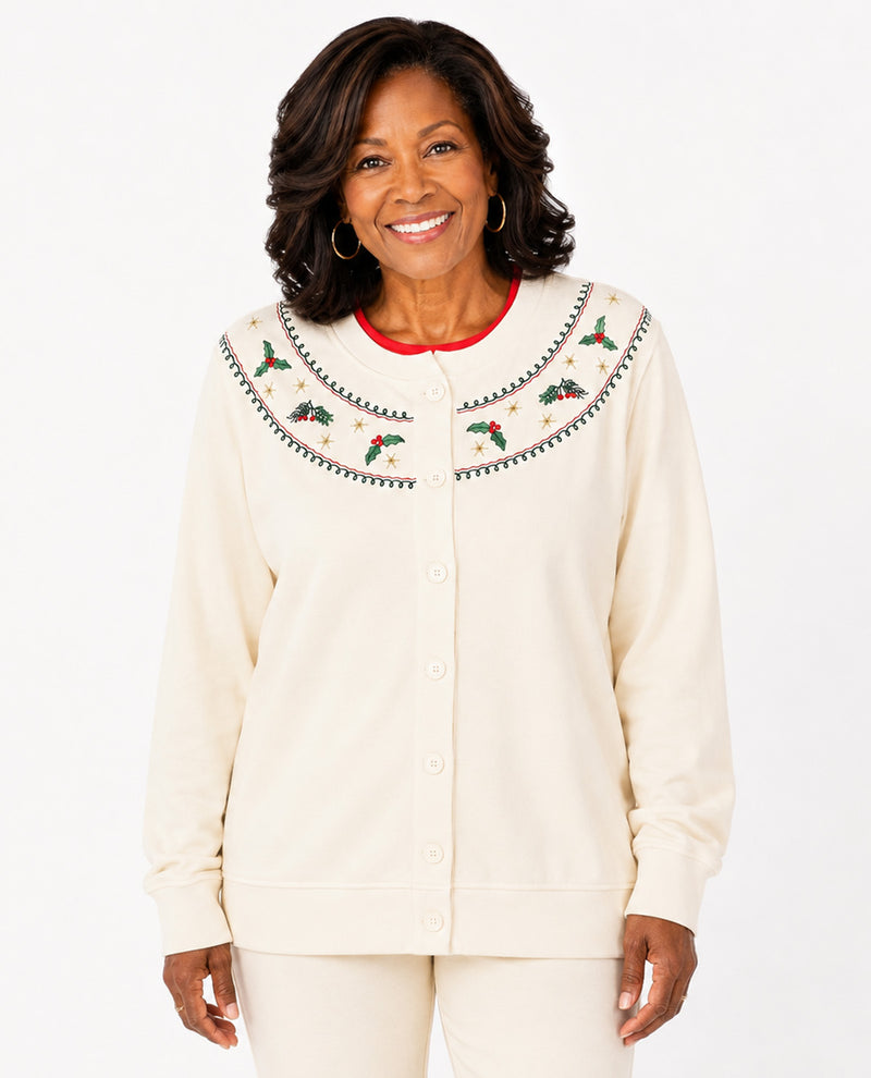A smiling woman wearing a cream-colored button-up cardigan with a large decorative yoke featuring embroidered holly leaves, pinecones, and red berries, paired with matching cream pants against a white studio background.